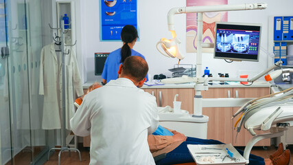 Elderly woman during the medical examination with male dentist in the dental office. Orthodontist speaking to patient sitting on stomatological chair while nurse preparing tools for surgery.