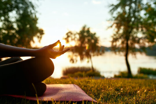 Unity with nature. Cropped shot of a woman doing yoga outdoors near lake or river at sunset, sitting in lotus pose and meditating on the background of nature
