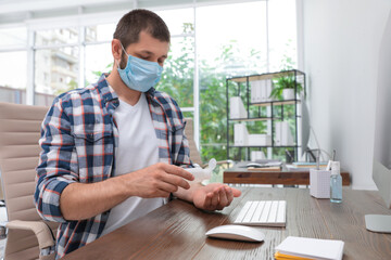 Office employee in mask applying hand sanitizer at workplace