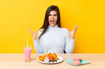 Young woman eating waffles and milkshake in a table over isolated yellow background unhappy and frustrated with something