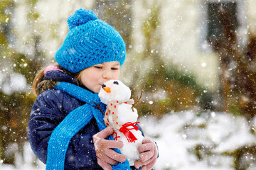 Cute little toddler girl making mini snowman and eating carrot nose. Adorable healthy happy child...