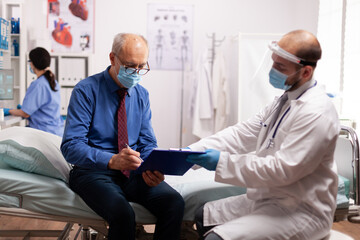 Elderly patient wearing face mask reading paperwork in the course of covid19. Healthcare medical specialist in clinic consultation room during global crisis with coronavirus.