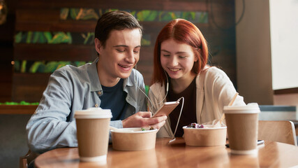 Happy teenage couple looking at the screen while watching something using smartphone and same pair of earphones, sitting in a cafe together on a daytime