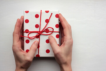 Person holding a gift box. Overhead view of female hands with present wrapped in red and white polka dot pattern paper on white background. Top view, copy space