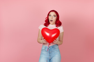 Fototapeta na wymiar Close-up a surprised, shocked young woman with red hair holds a large flying red heart-shaped balloon isolated on a pink background.