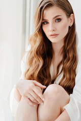 Naklejka premium beautiful woman in white robe sits on windowsill.morning of bride before wedding