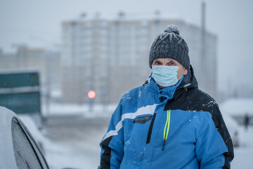 Fototapeta na wymiar Old man wearing knitted cap and protective face mask stands near car on roadside