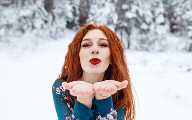 Young red-haired girl with long red hair on a winter background.Air kiss. Red-haired woman in a summer dress on the background of winter nature. - Powered by Adobe