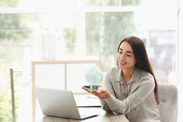 Young woman enjoying air flow from portable fan at workplace, space for text. Summer heat