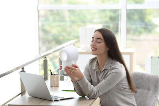 Young woman enjoying air flow from portable fan at workplace. Summer heat