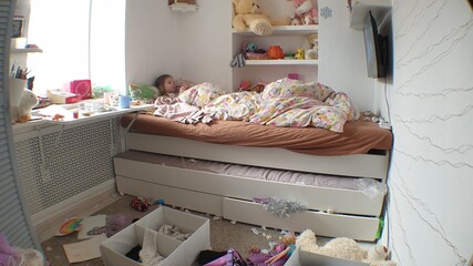clutter in the children's room, a little girl lies in bed and watches TV in an untidy room