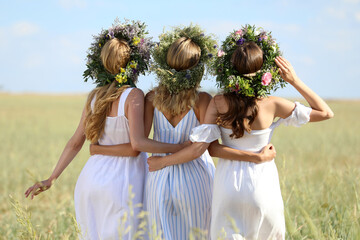 Young women wearing wreaths made of beautiful flowers in field on sunny day, back view