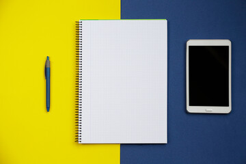 Top view of an open notebook, a tablet pc, and a pen against blue and yellow background.