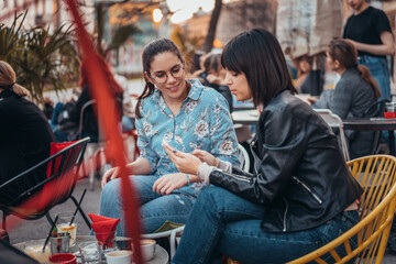 Fototapeta na wymiar Two beautiful women in cafe outdoors looking at something on mobile phone. Two female using mobile phone application shopping online and enjoying coffee outside in city