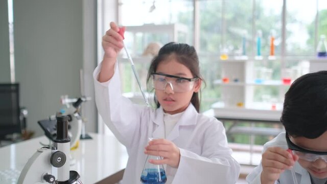 Young scientist girl with eye protecting glasses use dropper to mix color chemical together and work near her classmate in classroom or laboratory with glass windows.