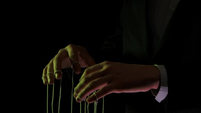 Puppeteer manipulating and pulling strings of marionettes. Male hands close up painted with colored lights on black background. People are toys in hands of dominant businessman dictator. Slow motion.