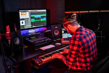 Hipster man with beard in headphone is playing keyboard and sining his new song in stereo studio to record brand-new track .