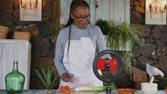 African senior woman preparing food recipe while streaming online with mobile smartphone cam for web kitchen masterclass channel