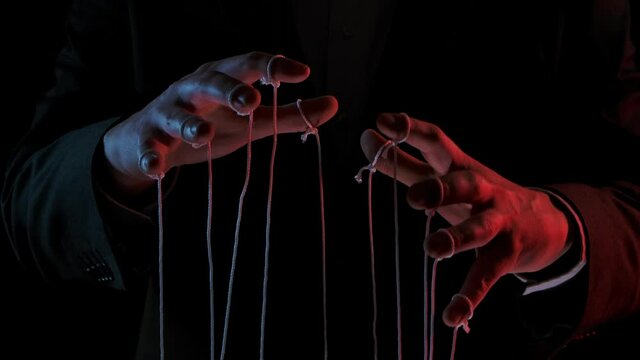 Puppeteer manipulating and pulling strings of marionettes. Male hands close up painted with colored lights on black background. People are toys in hands of dominant businessman dictator. Slow motion.
