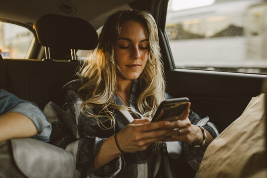 young caucasian woman using smartphone sitting in backseat of car