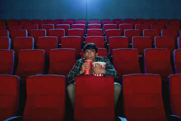 Focused young man holding a drink and popcorn basket while watching movie alone in empty theater auditorium