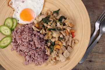 Thai fusion stair fried chicken with Thai basil and cashew nut with health brown rice fried egg sauce cucumber on woody plate over rustic wood background fork spoon