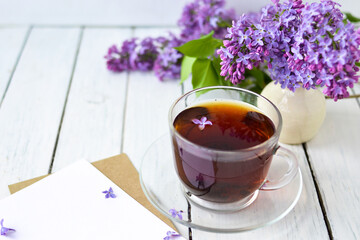Delicate morning tea table setting with lilac flowers, a transparent cup of tea and a saucer and a white vase, white and craft note paper on a white wooden board. Mockup. Copy space. 