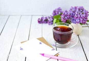 Delicate morning tea table setting with lilac flowers, a transparent cup of tea and a saucer and a white vase, white and craft paper for notes, and a pink pen on a white wooden table. Mockup. 