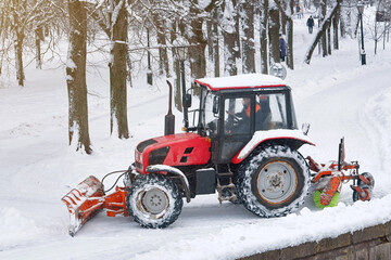 Snow removal tractor with scoop and rotating brush sweeping snow from footpath in park. Red tractor automated rotating brush removes snow from walkway in city park. Municipal road sweeping vehicle