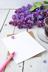 Delicate morning tea table setting with lilac flowers, a transparent cup of tea and a saucer and a white vase, white and craft paper for notes, and a pink pen on a white wooden table. Mockup. Copy