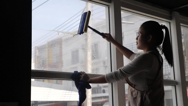 Home routine, cleanliness concept. Asian woman clean a window pane with a squeegee and soap suds foam. Hands in blue protective gloves cleaning glass on the windows with a detergent spray.