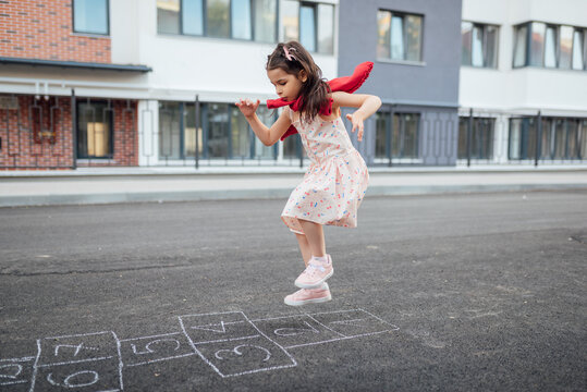Full view of little girl playing hopscotch with her mother on playground outdoors. Horizontal image of a child plays outside. Kid plays hopscotch drawn on pavement. Activities for kids outside.