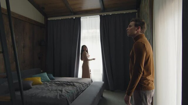 Handheld slowmo shot of smart asian woman opening curtains in luxurious hotel room spending vacation there with her boyfriend, looking together out of window