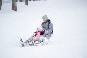 Happy young adult father with baby girl sitting on sledge and sledding down on snow from hill. Enjoying white winter day at park. Spending time together in weekend. Side view.