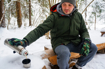 a guy in a winter forest drinks a hot drink from a thermos bottle, a tourism concept