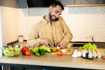 Fototapeta premium Young man is talking on phone while cooking salad in the kitchen. Vegan guy is cutting vegetables for the salad.