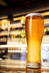 Nightlife. Full glass of light lager beer with drops on the side, wooden table in warm light of...
