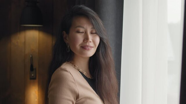 Medium slowmo close-up portrait of beautiful smart asian woman standing by window in dark hotel room smiling to camera