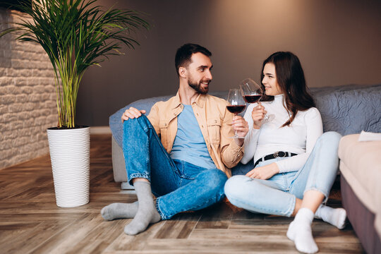 Young couple is sitting on the floor with red wine in bedroom and celebrating their new apartment.