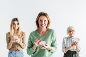 cheerful young woman standing with smartphone near mother and young daughter on blurred background isolated on white