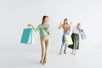 Naklejka premium full length of happy woman with shopping bags near daughter and senior mother on blurred white background
