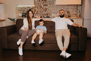 Obraz premium Dad, son, and mom are watching boring TV programs on the sofa in the apartment. The family is enjoying the evening. Relatives at home. A young mother with long hair is smiling.