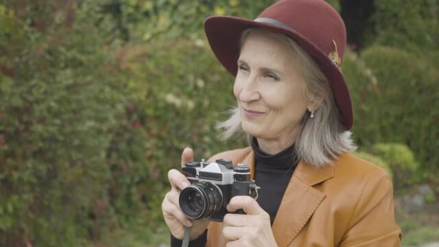 Close-up side view of positive creative senior woman taking photos on camera in spring or autumn park. Portrait of cheerful happy Caucasian retiree enjoying nature outdoors. Hobby and lifestyle.