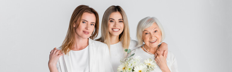 three generation of happy women smiling while hugging near flowers isolated on grey, banner