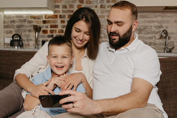 A dad, a son, and a mom are watching video on the sofa. A happy husband with a beard is demonstrating the show on the smartphone to a laughing child and a wife in the evening at home.