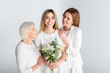 cheerful young woman smiling while holding flowers near mother and granny isolated on grey