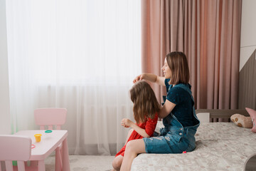 Happy delightful aunt wearing comfy jeans romper, spend time with her cute lovely niece, combing...