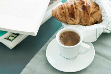 Morning coffee concept, white cup of coffee, empty book page, fresh baked croussant in paper pack on gray cloth and dark grey background