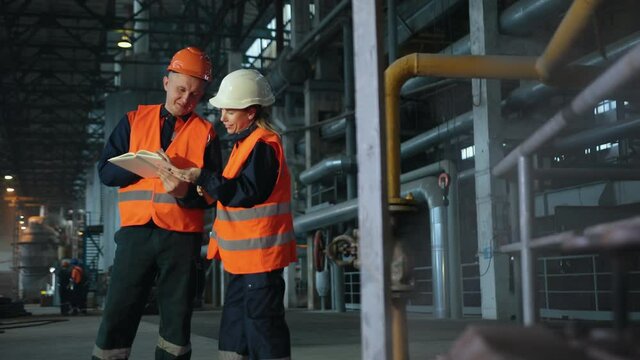 Two maintenance engineers man, woman inspect. Work heavy industry manufacturing factory. Mechanic, female engineer talking in plant. Young employees wearing helmets are working industrial factories