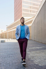 Portrait of a young bearded man, model of fashion, in urban background wearing casual clothes while walking with hands on pocket